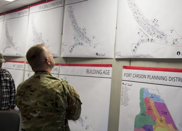 Military personnel examines wall maps titled "Building Conditions" and "Fort Carson Planning District" in an office setting