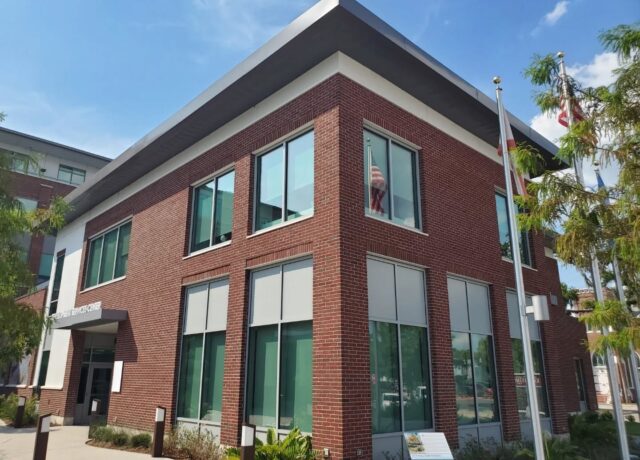 Red brick building with large windows and flags outside, against a clear blue sky.
