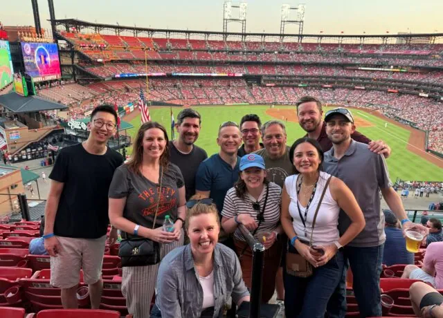 Group of ten people smiling, posing at a baseball stadium, with a crowded field in the background