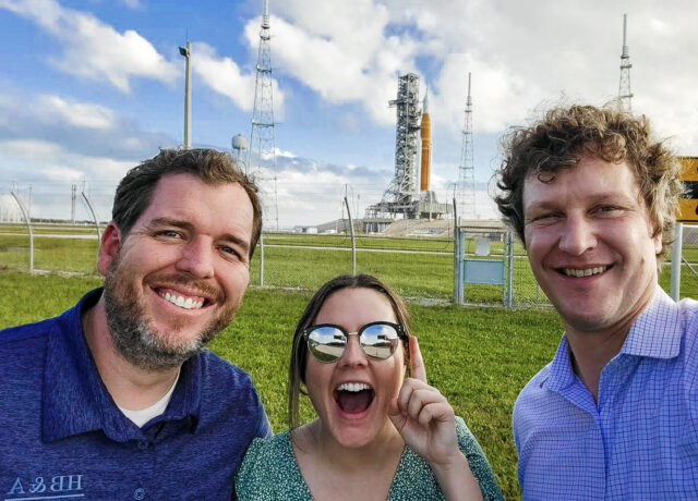 Three smiling people pose with excitement near a launch pad