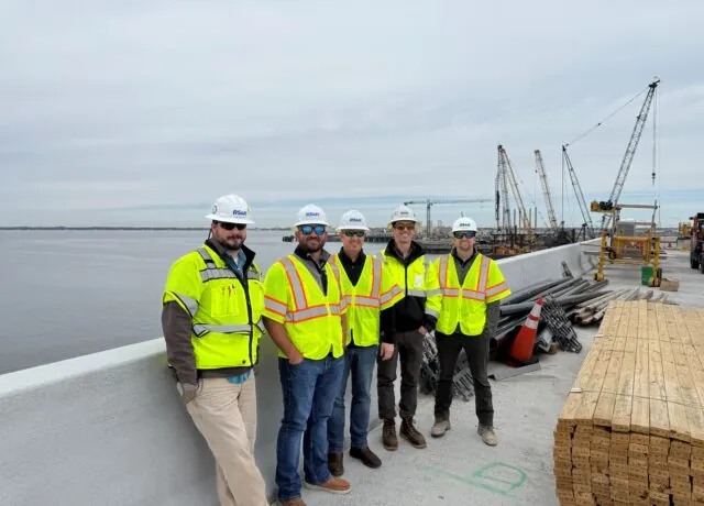 Brent Wilkins and team on bridge construction site, wearing safety vests and helmets, with cranes in the background