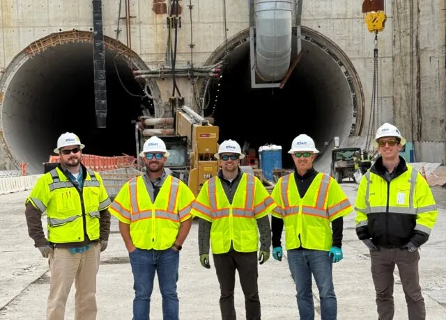 Five construction workers in safety gear stand in front of twin tunnel entrances at a worksite