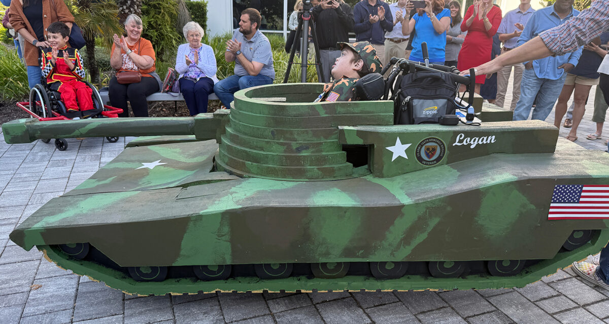 Child in wheelchair dressed in camouflage tank costume named "Logan," surrounded by applauding people. American flag visible