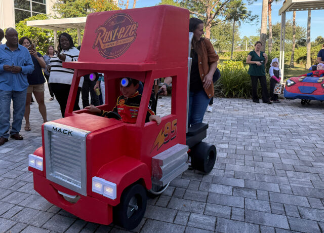 Child in red truck costume with "Rust-eze" logo, assisted by an adult, surrounded by onlookers on a paved area