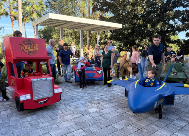 Children in creative vehicle costumes, including a red truck and blue plane, surrounded by adults outdoors