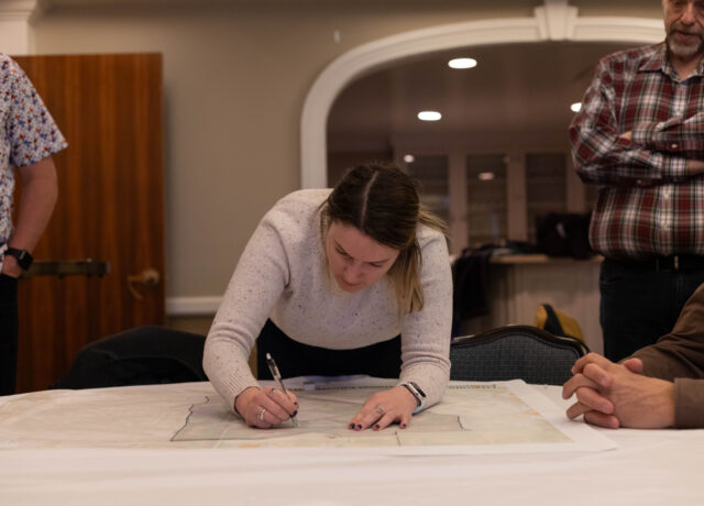 Person leaning over table, intensely writing on a large map or document, surrounded by others observing