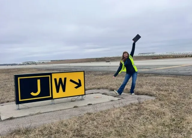 Julie enthusiastically poses next to an airport taxiway sign marked "J W."