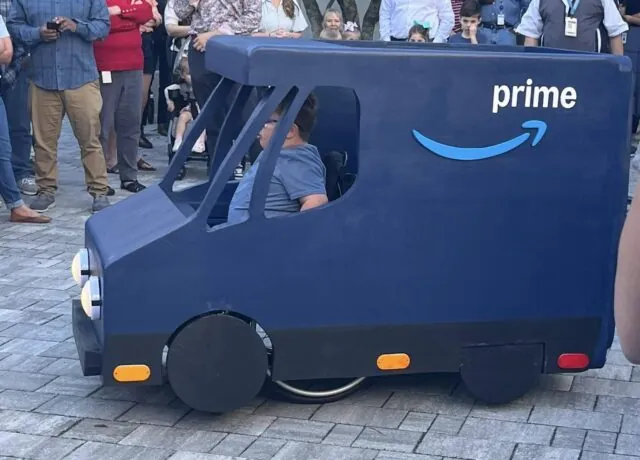Person in a small, blue truck costume resembling an Amazon Prime vehicle, with crowd watching
