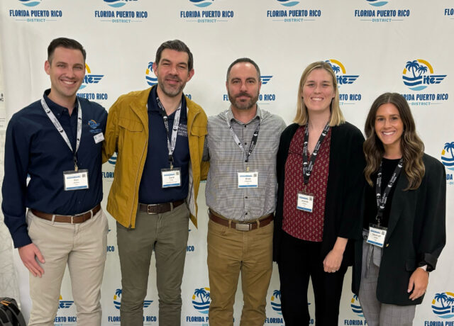Five people standing, smiling, in front of a "Florida Puerto Rico District" backdrop, wearing name tags