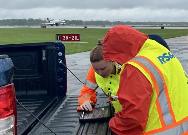Two individuals in reflective gear work on a tablet in a truck bed at a rainy airport runway