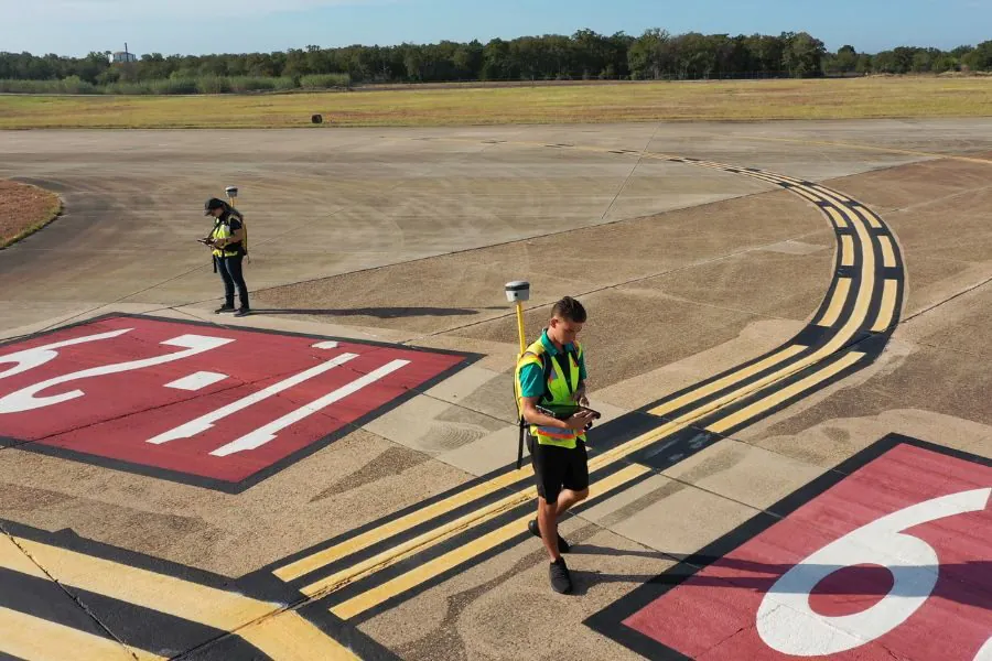 Two workers in safety vests surveying airport tarmac with equipment