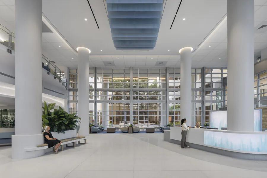 Modern lobby with high ceilings, columns, and large windows; one person sitting, another at the reception desk