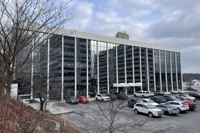 Modern office building with glass facade, parked cars, and overcast sky.