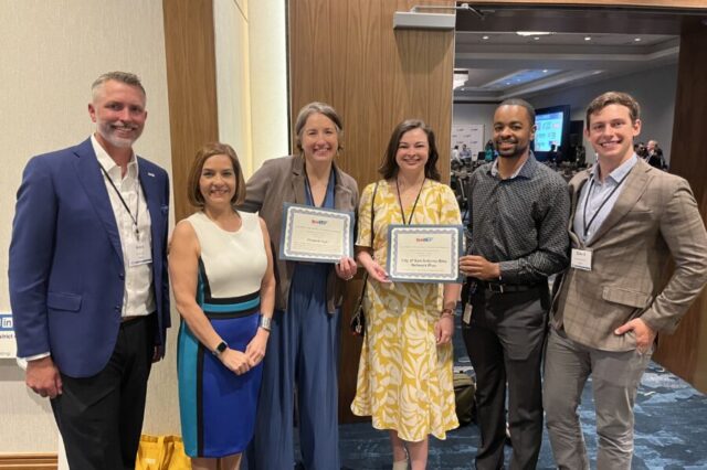 Six people standing, two holding certificates, smiling at a conference.