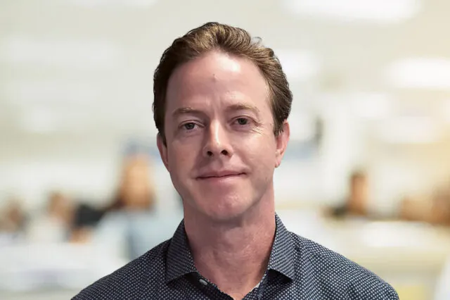 Mattos Mason smiling in an office setting, wearing a dark patterned shirt