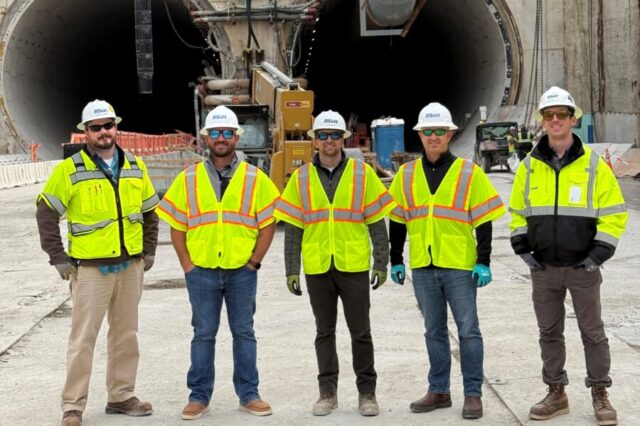 Five construction workers in safety gear stand in front of a tunnel entrance