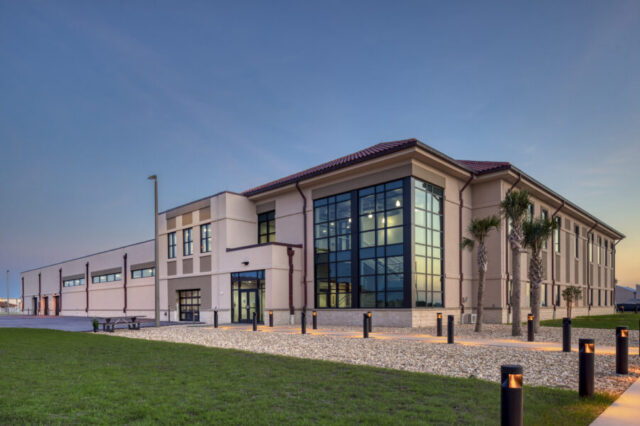 Modern building exterior with large glass windows at dusk, surrounded by palm trees and illuminated pathways