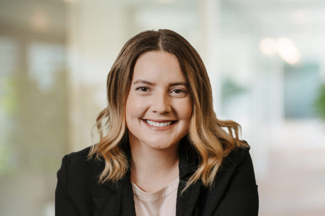 Tara smiling warmly, wearing a black blazer, in a softly blurred office background
