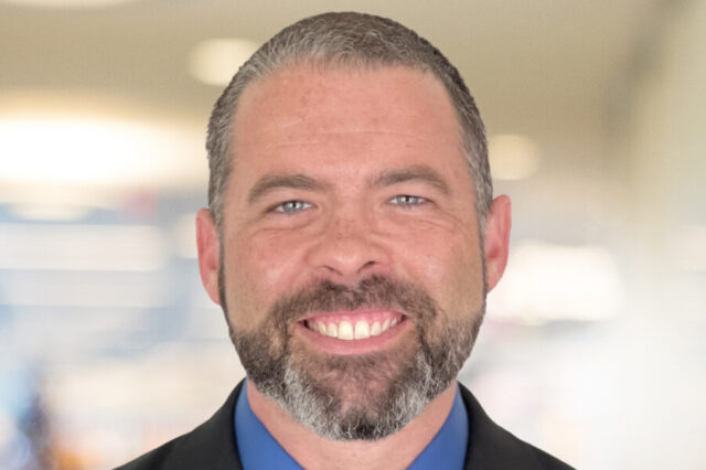 Chris Duguid smiling in a professional setting with a blurred background