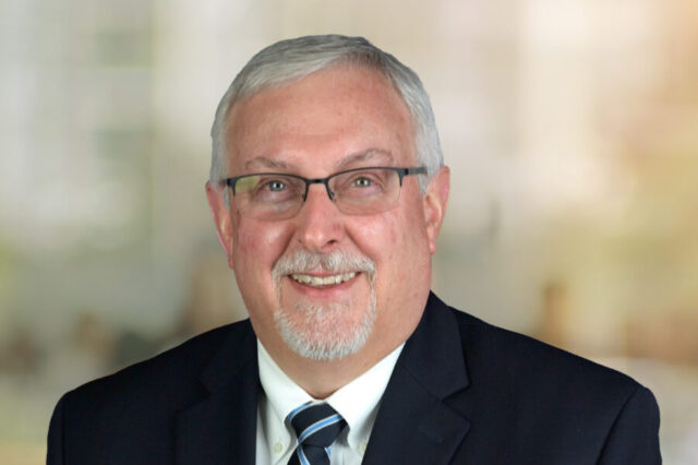 Everett Tom smiling in a suit with glasses, neutral background