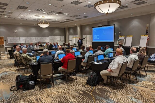 People seated in conference room, viewing presentation on large screen with instructor speaking, charts and notes on walls