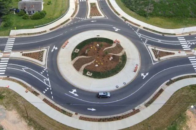 Aerial view of a roundabout with a central garden, road markings, and a car driving around it.