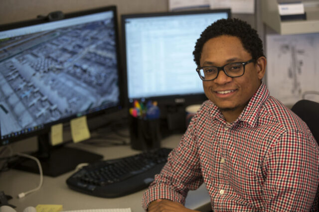 Gareth Hanley smiling at desk with dual monitors displaying maps and data.