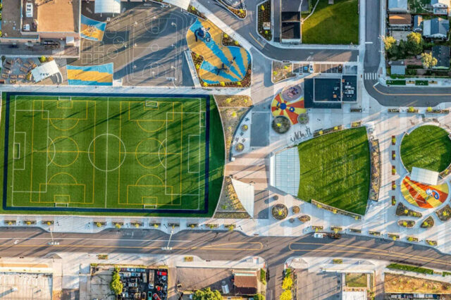 Aerial view of a park with a soccer field, colorful playgrounds, and landscaped green spaces surrounded by urban development.