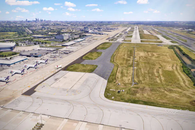 Aerial view of airport runway and taxiway with parked airplanes, city skyline visible in the distance.