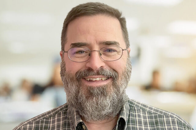 Lee Tim smiling, wearing glasses and a plaid shirt, blurred background.