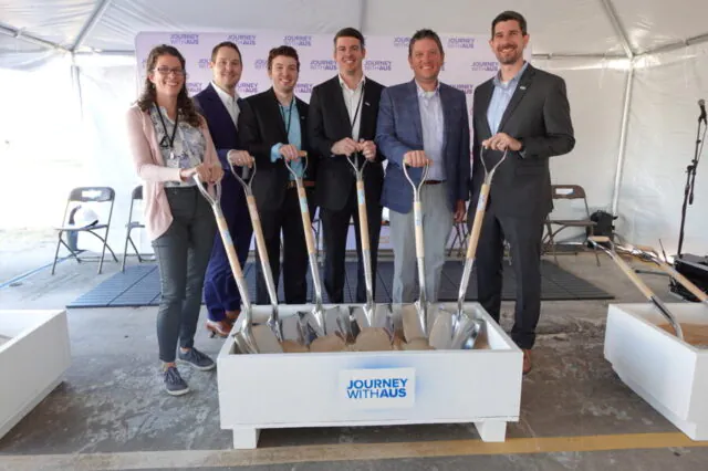 Six people with shovels at a groundbreaking event, banner reads "Journey With Aus"